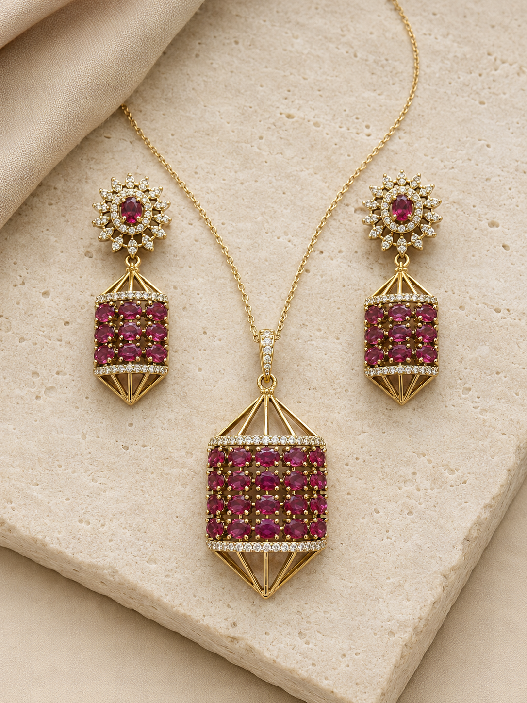 Gold jewelry set with a pendant necklace and matching drop earrings, featuring deep pinkish-red gemstones and small clear stones, displayed on a beige stone surface.