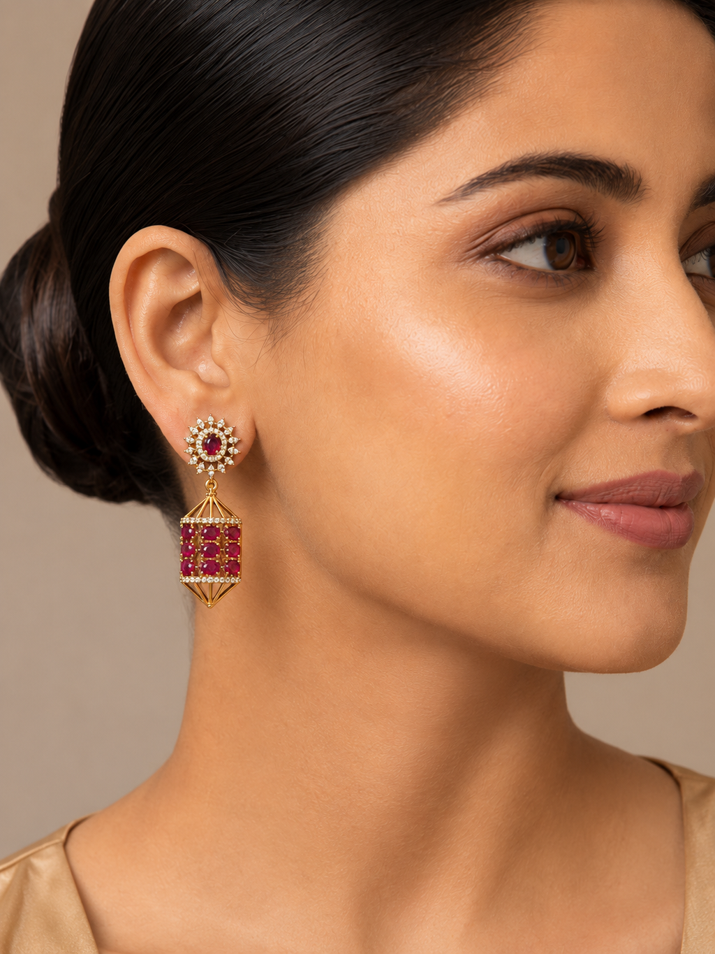 A close-up of a woman wearing a gold-toned dangle earring with a round red stone top and a faceted rectangular red stone drop, shown against a soft beige background.